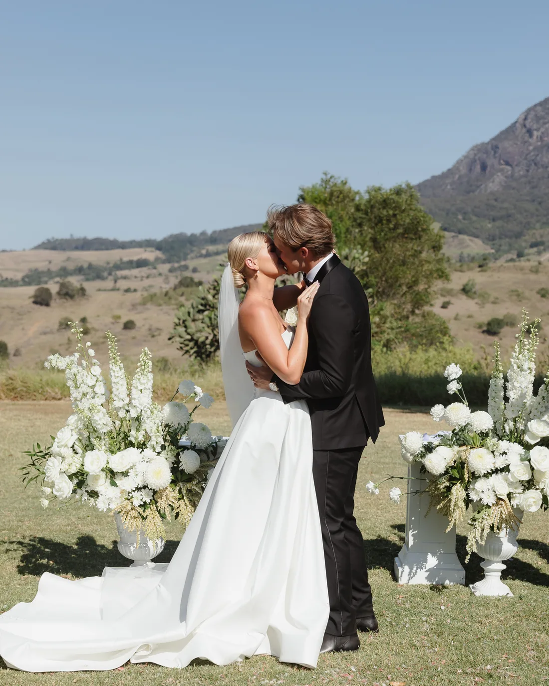 Alaska and Billy kissing at the altar, white floral urns and mountain views behind