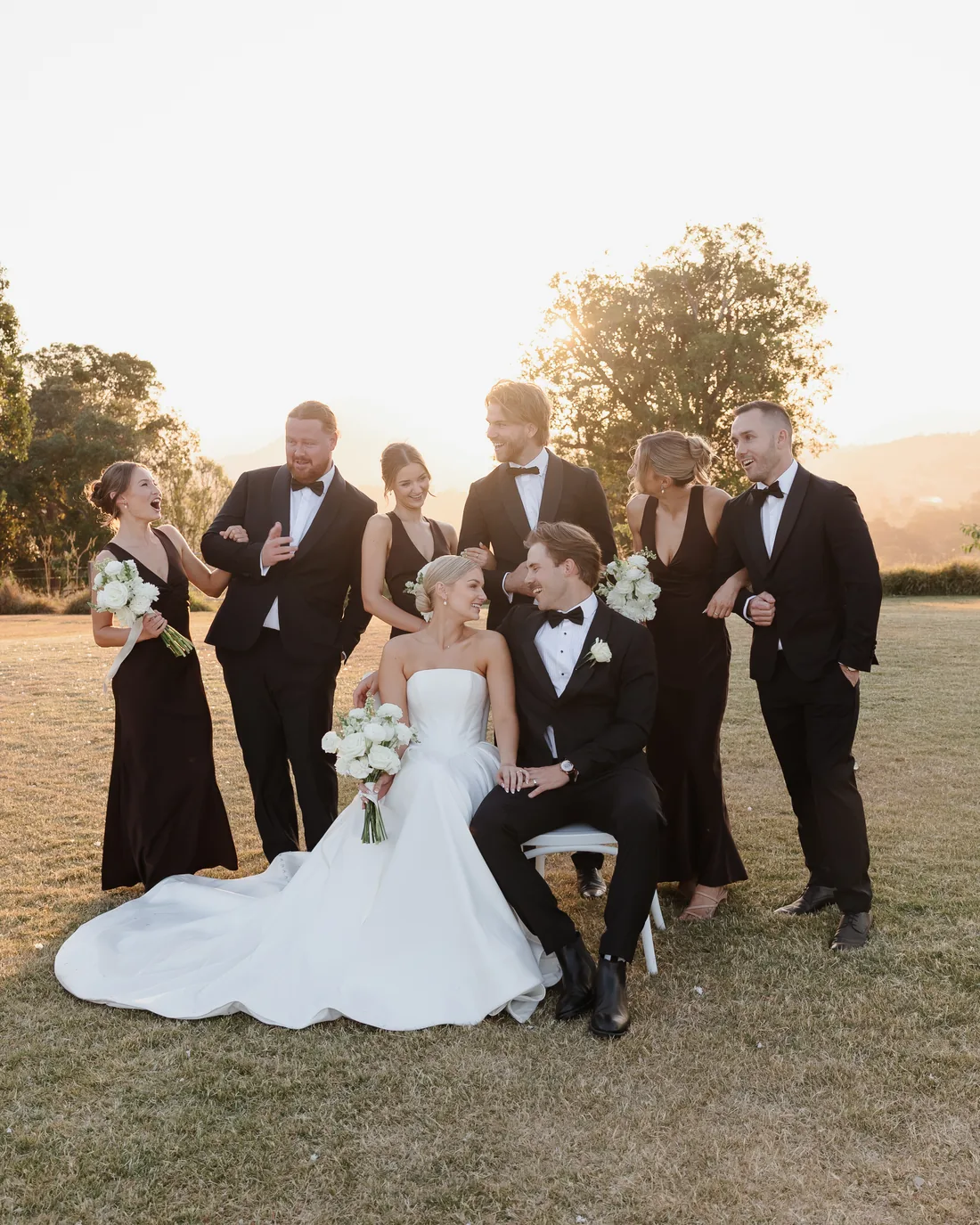 Full bridal party laughing together at sunset, bride and groom seated centre