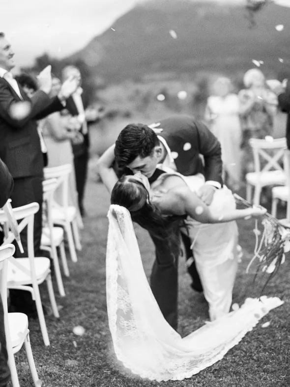 Black and white photo of Will dipping Alex for a confetti kiss at the end of the aisle