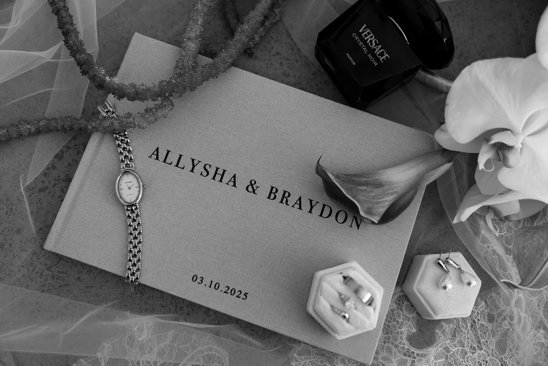 Black and white flatlay of the Ally and Braydon wedding invitation, watch, rings and orchid florals
