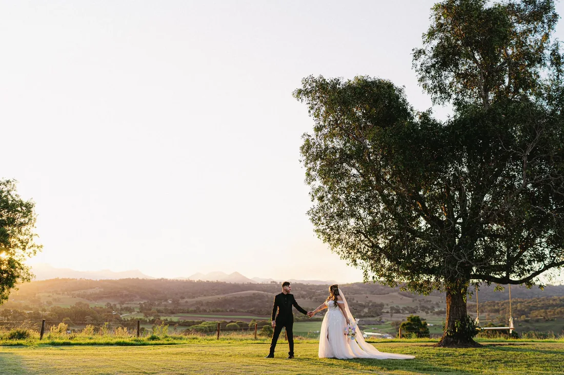 Chelsea and Rhys walking hand in hand beneath a large tree at golden hour