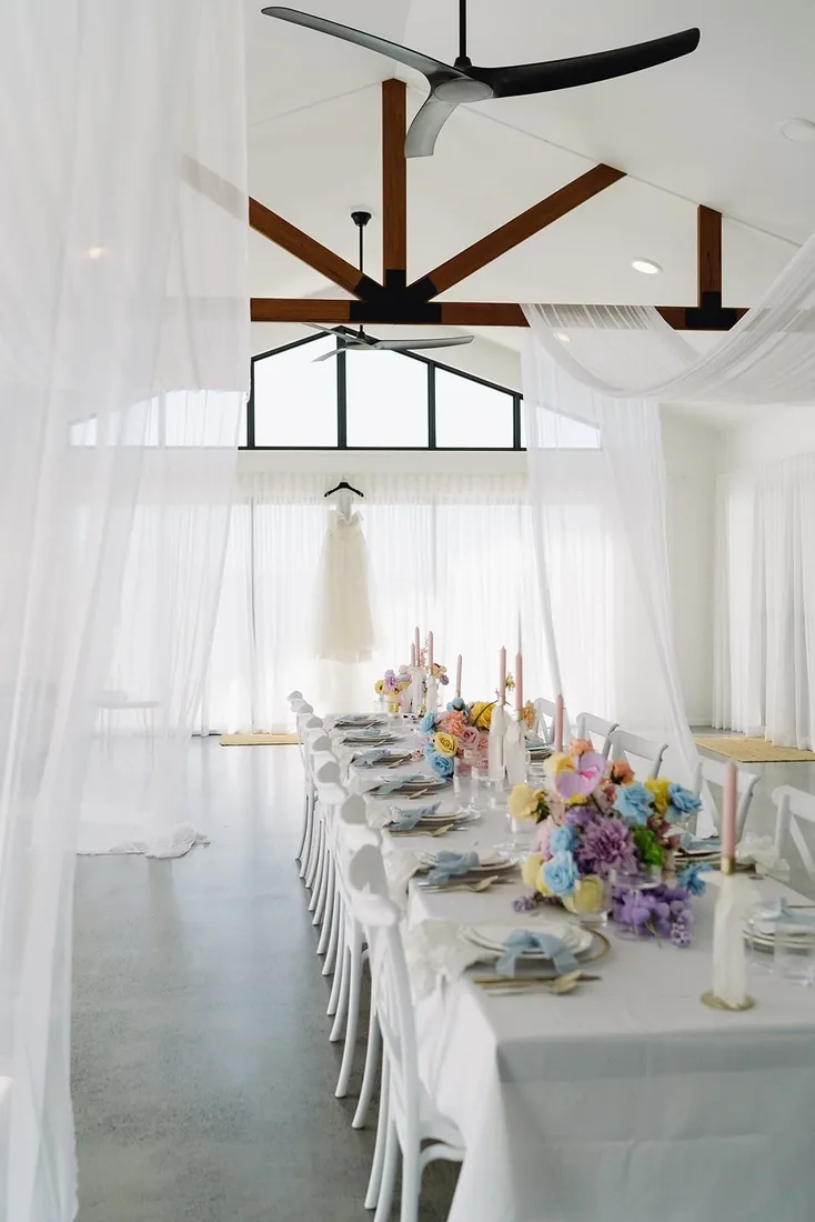 Reception room with sheer draping and timber beams, wedding dress hanging above a long table set with pastel florals
