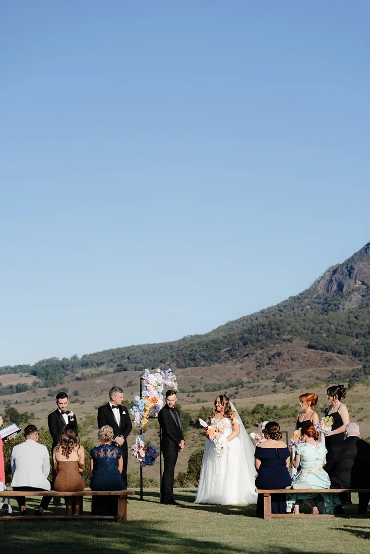 Wedding ceremony in progress with the celebrant and bridal party, mountains behind and guests seated on timber benches