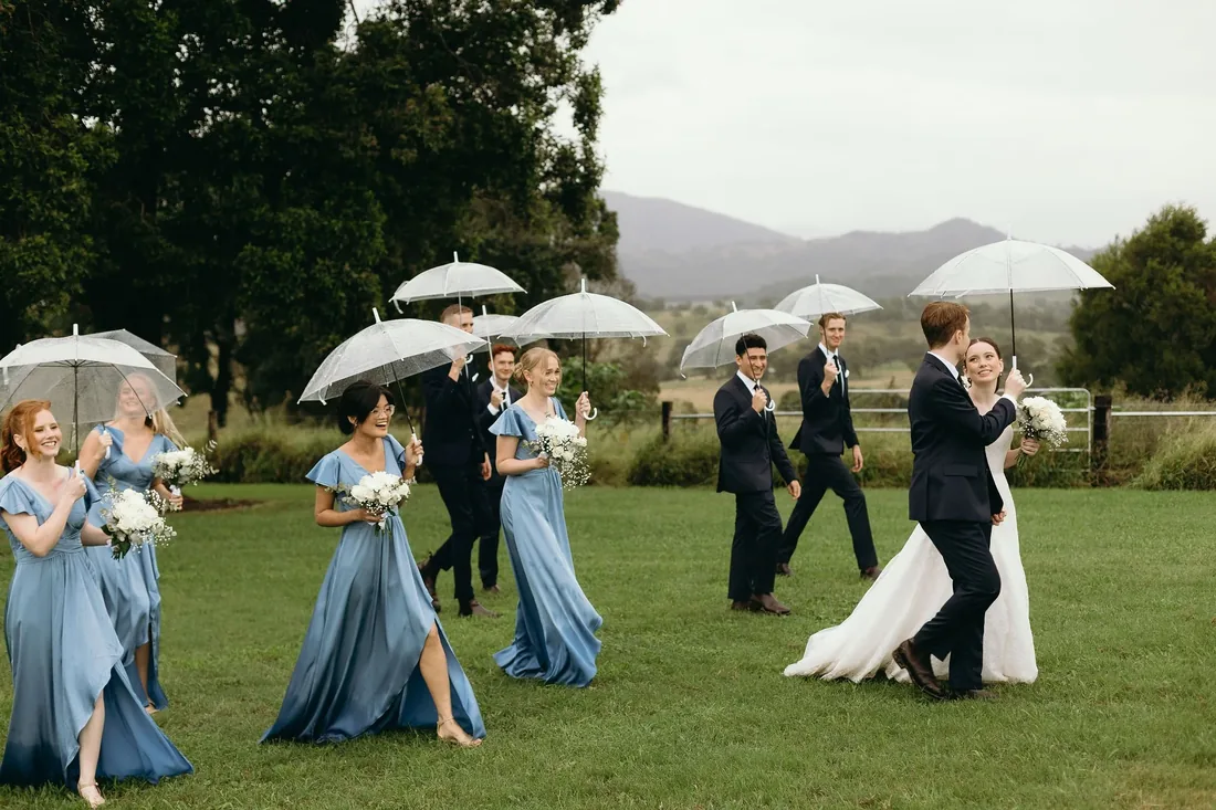 Bridal party walking across the lawn holding clear umbrellas in the rain, bridesmaids in dusty blue