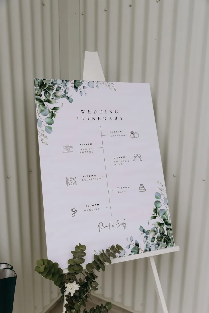 Wedding itinerary sign styled with eucalyptus leaves against a corrugated iron wall