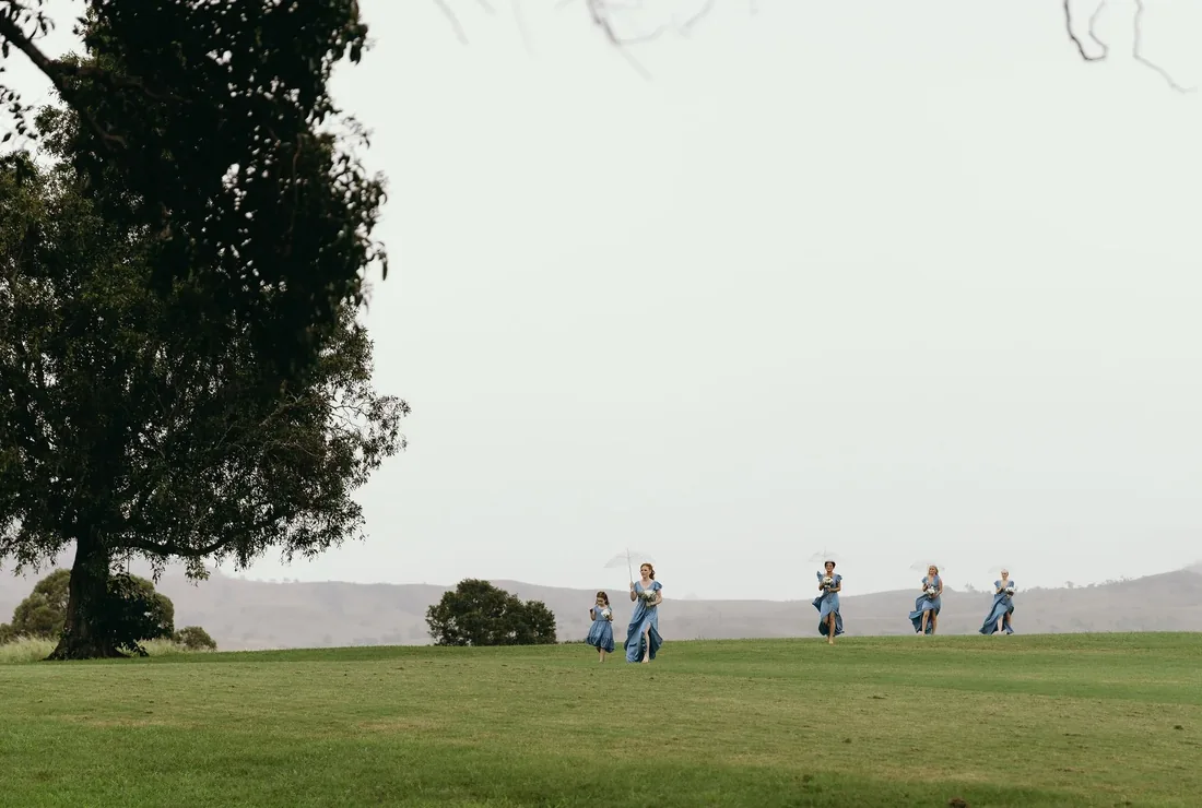 Bridesmaids in dusty blue dresses running across the lawn holding bouquets