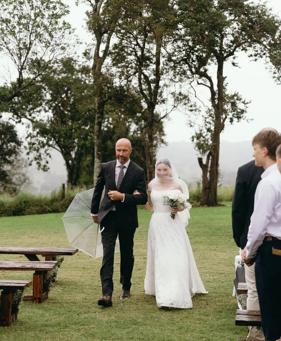 Emily's father walking her down the aisle under an umbrella, guests seated on timber benches