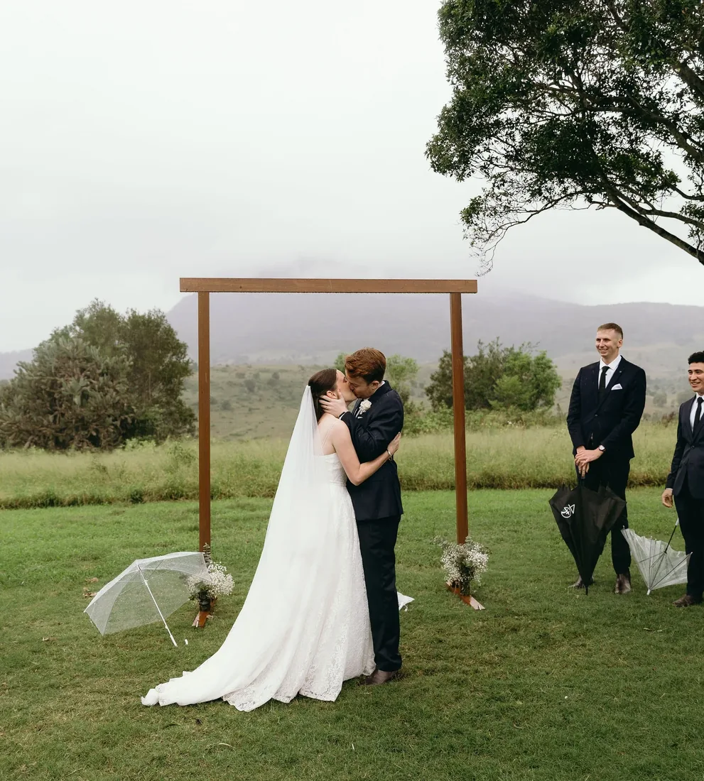 Daniel and Emily kissing at the timber ceremony arch in the rain, umbrellas set either side