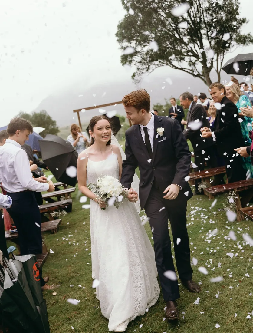 Daniel and Emily walking back down the aisle through confetti as guests cheer under umbrellas