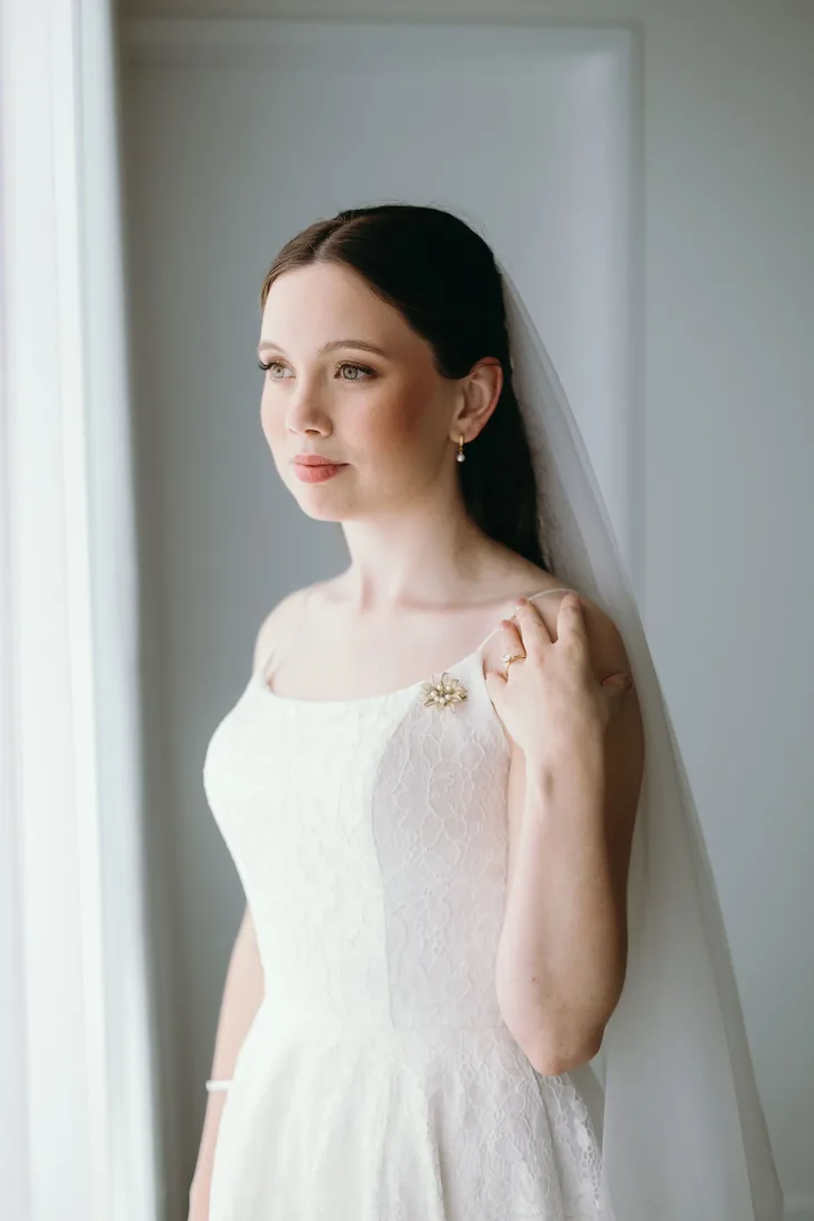 Emily's bridal portrait by a window, wearing her veil and a brooch pinned to her lace gown