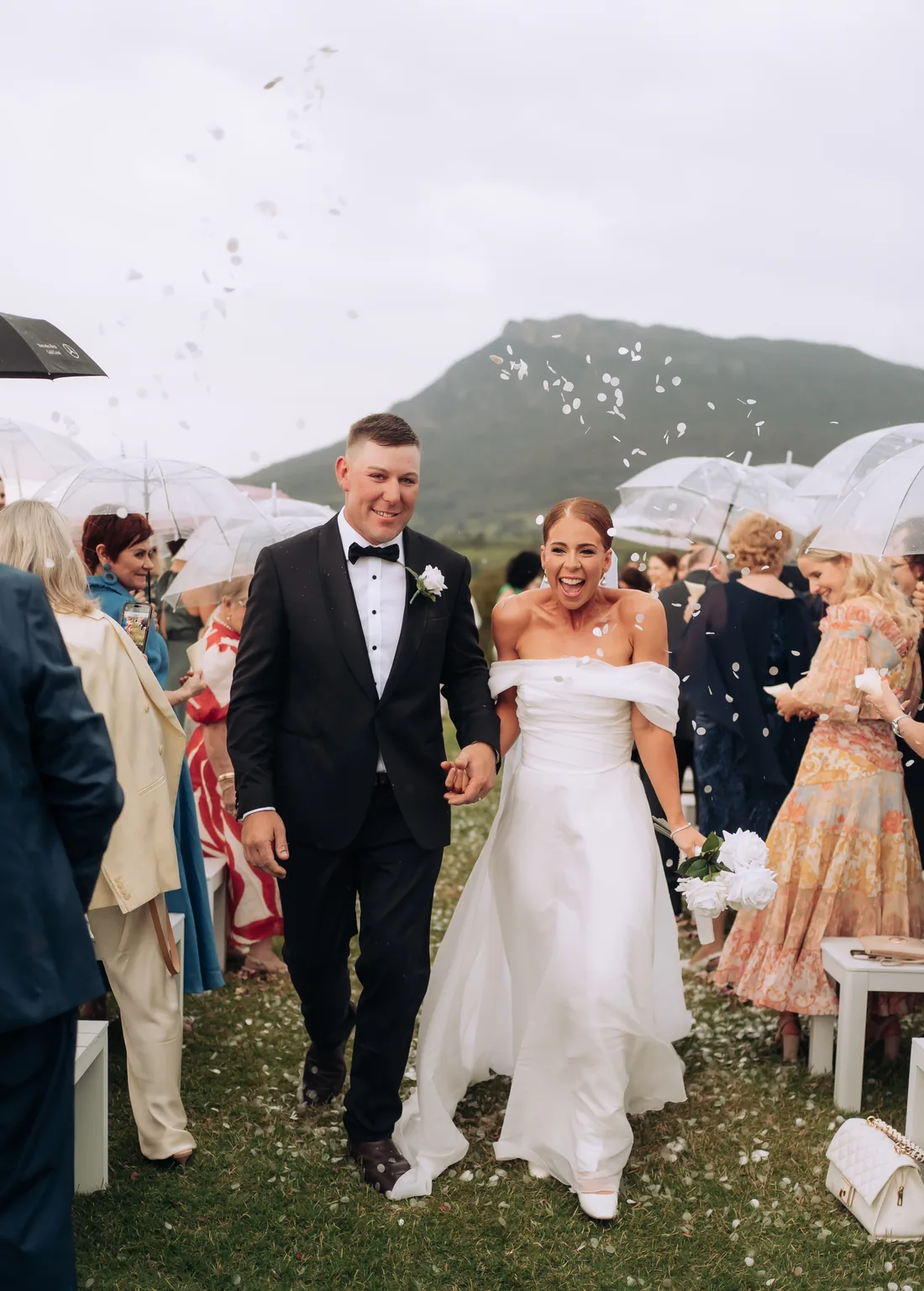 Deanna and Michael walking through confetti under clear umbrellas in the rain