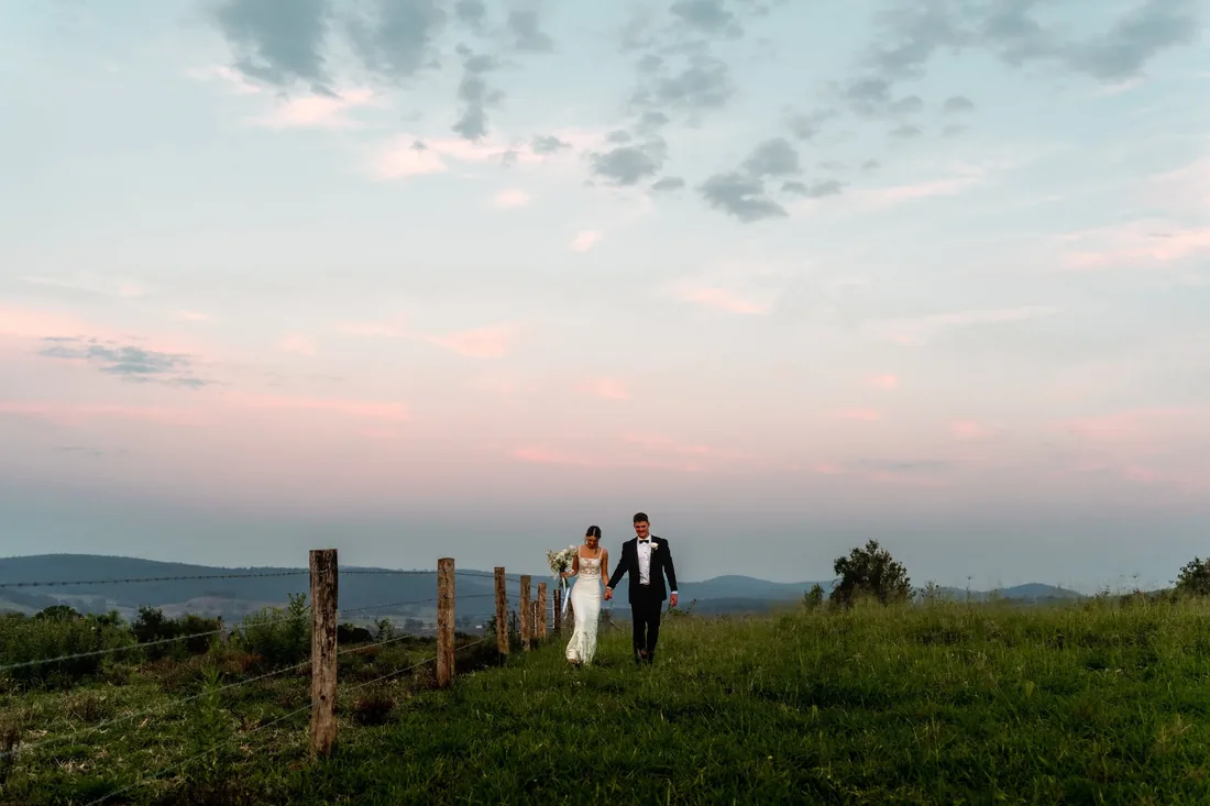 Georgia and Nick walking hand in hand along a fence line at dusk, pastel sky overhead