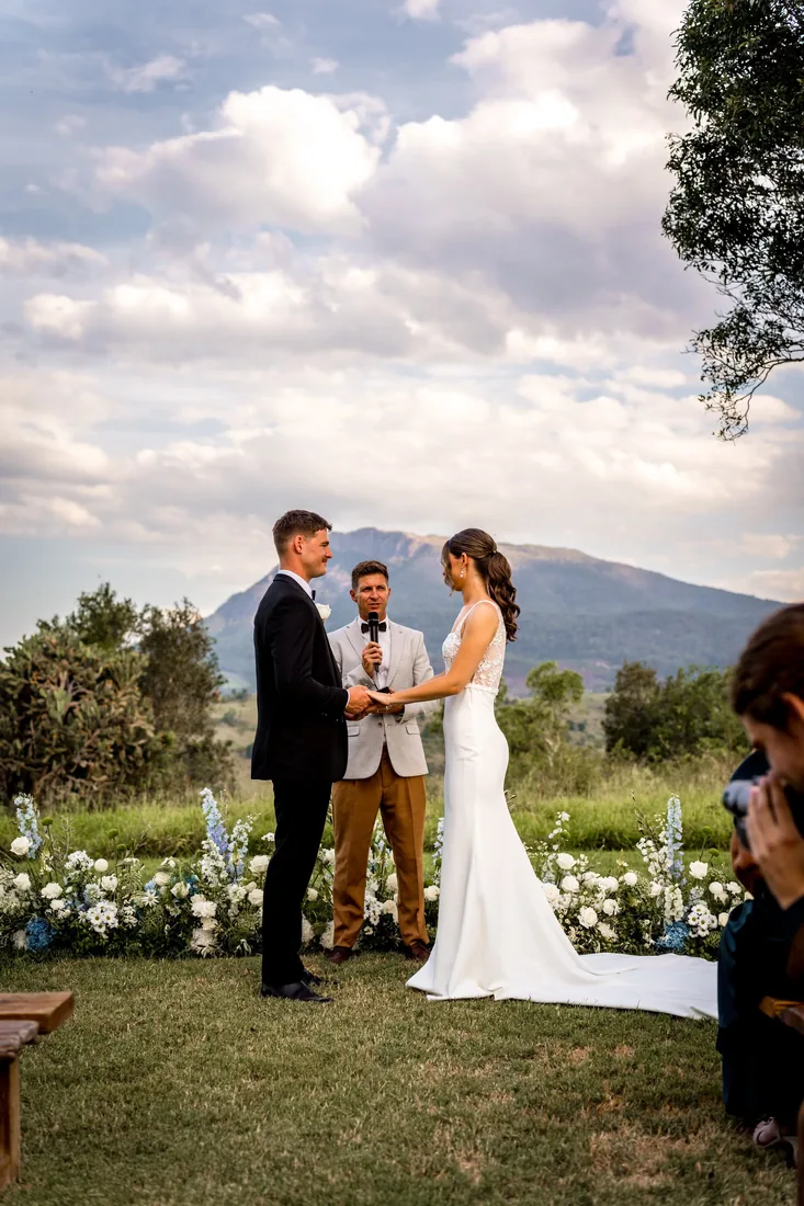Georgia and Nick holding hands during their ceremony, celebrant beside them and blue and white floral arrangements either side