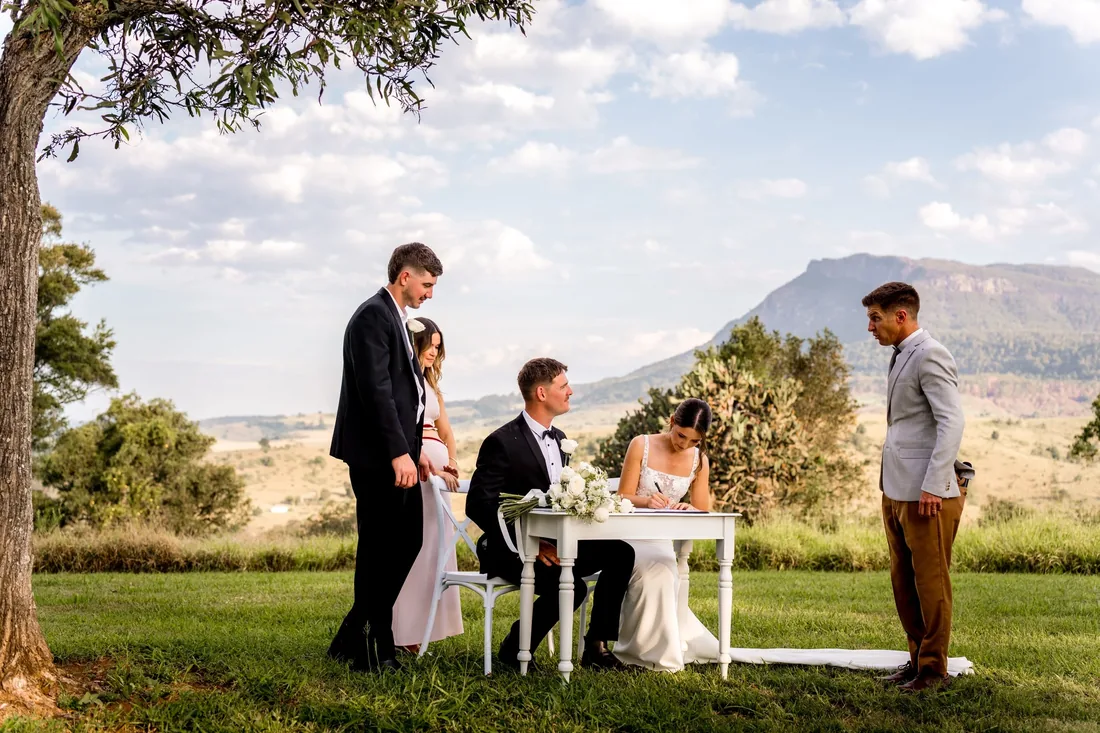Signing the wedding register at a table on the lawn as guests and the celebrant look on, mountain views behind