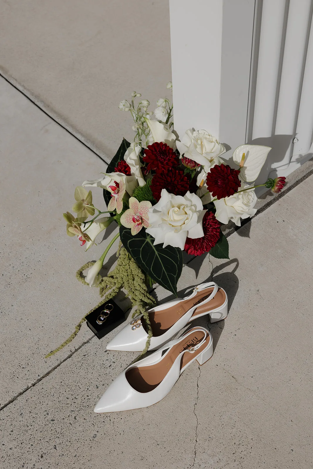 Bridal bouquet of white roses and burgundy dahlias styled beside white heels and rings