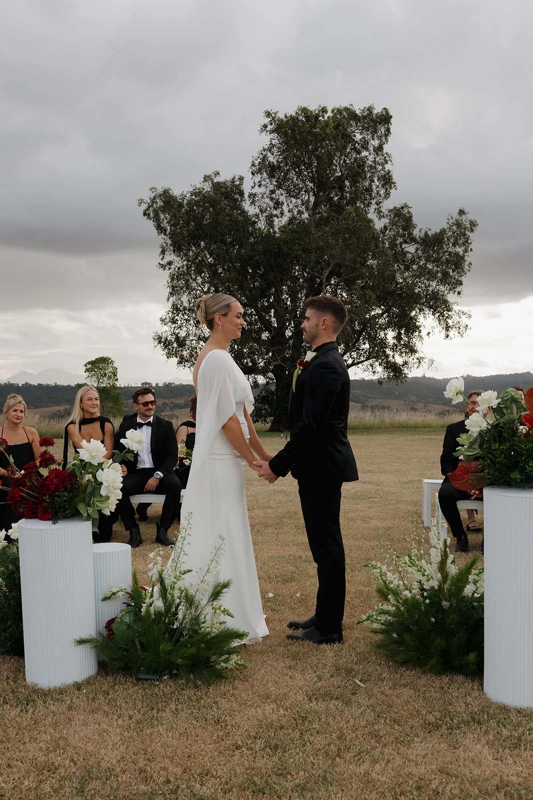 Jess and Cory holding hands during their vows beneath a large tree