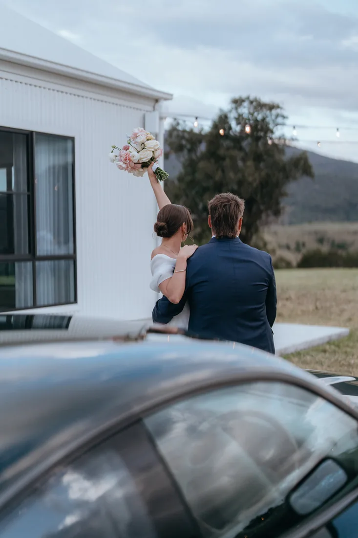 Jorjia and James walking away from the venue building, Jorjia holding her bouquet in the air