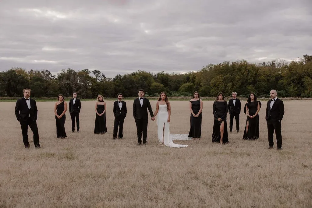 Full bridal party dressed in black standing in formation in a golden field with the bride in white at the centre