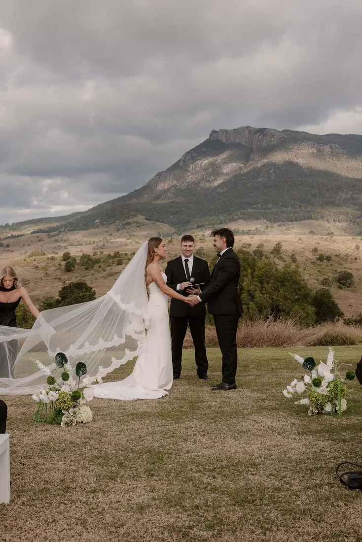 Karly and Anru holding hands during their vows, celebrant and best man beside them, mountains behind