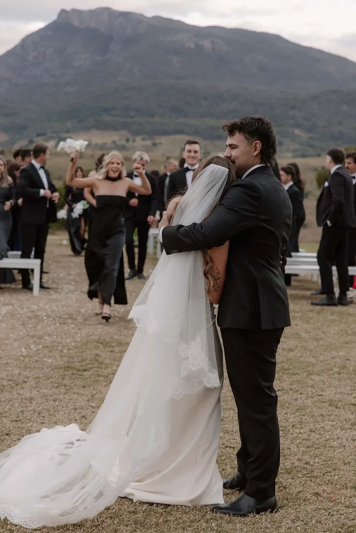 Karly and Anru embracing after their ceremony as the bridal party celebrates behind them