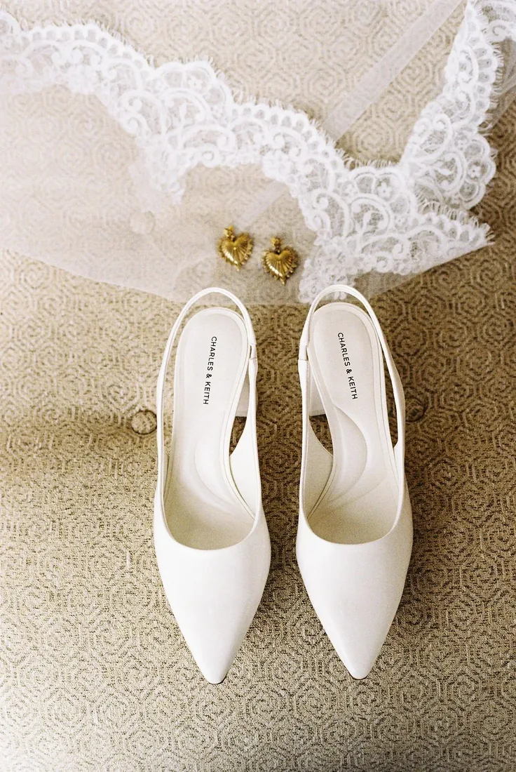 Bridal shoes, gold heart earrings and lace veil detail styled on a textured rug