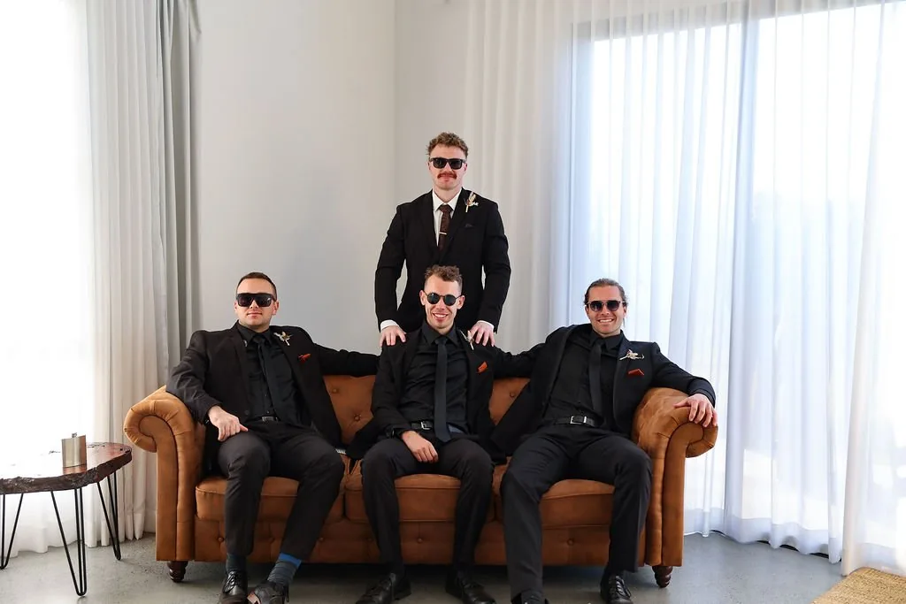 Four groomsmen in black suits and sunglasses posing on a leather couch