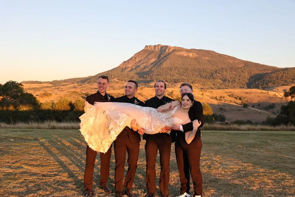 Groomsmen carrying Kate across the field at sunset with Mount Maroon behind