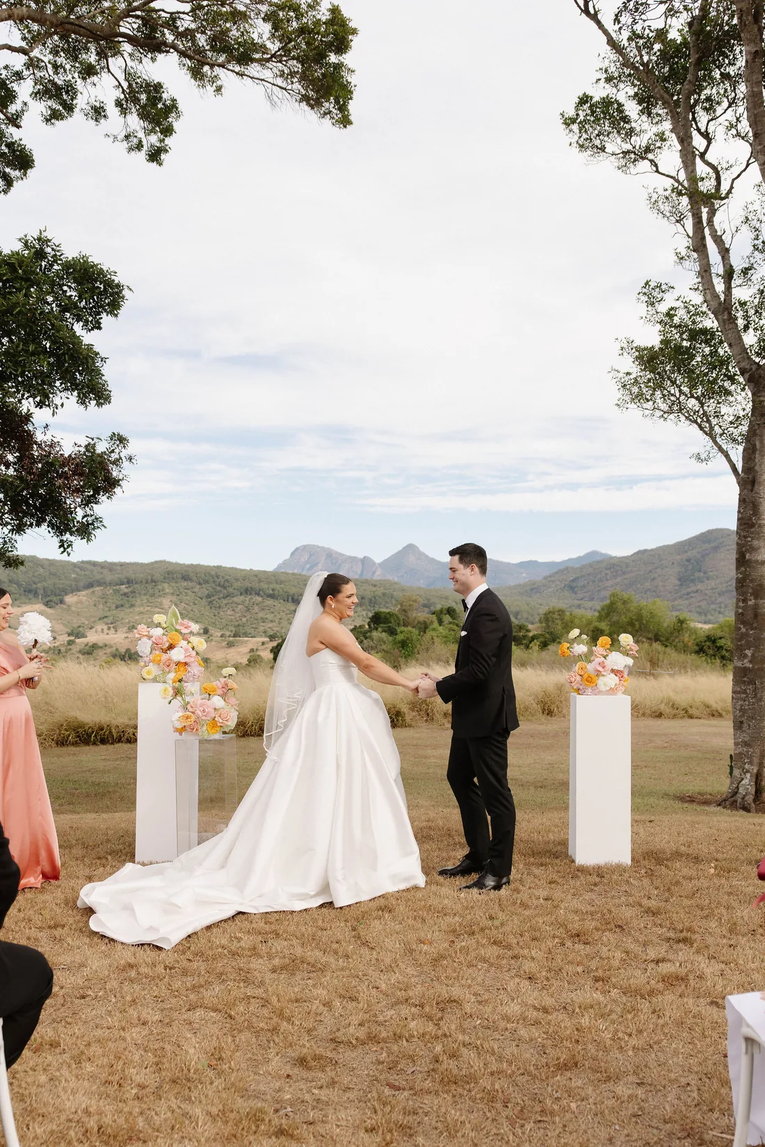 Laura and Connor holding hands during their vows, floral pillars and mountains behind