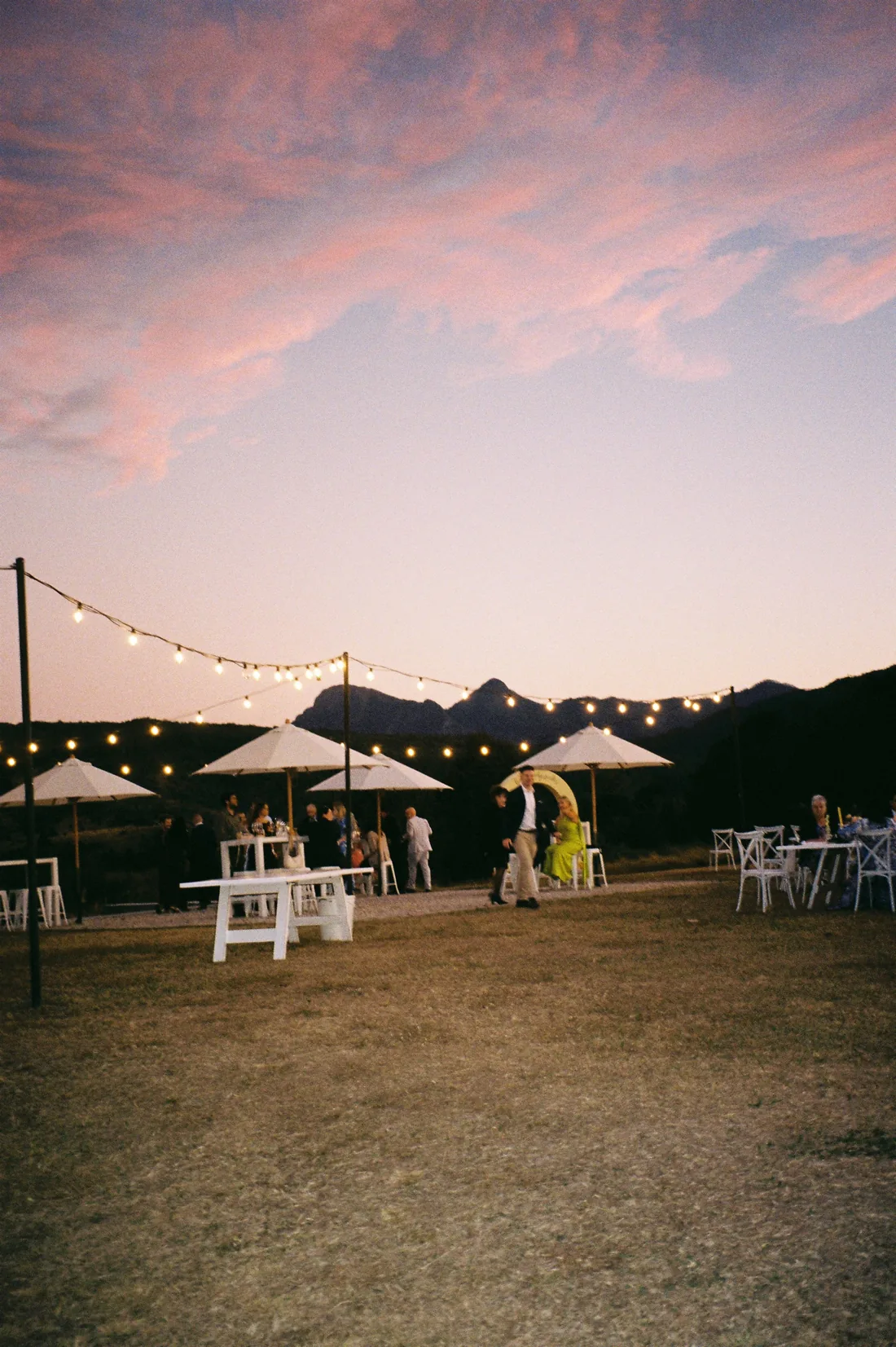 Guests mingling at dusk under festoon lights and market umbrellas, mountains silhouetted behind