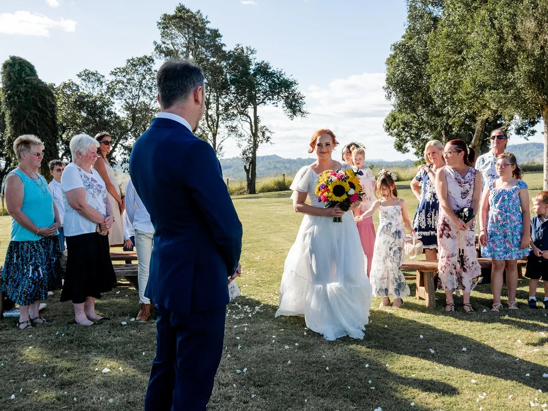 Russell watching Leisa walk down the aisle carrying a sunflower bouquet, flower girl beside her