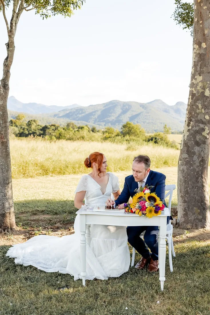 Leisa and Russell signing the register at a table on the lawn, mountains in the background