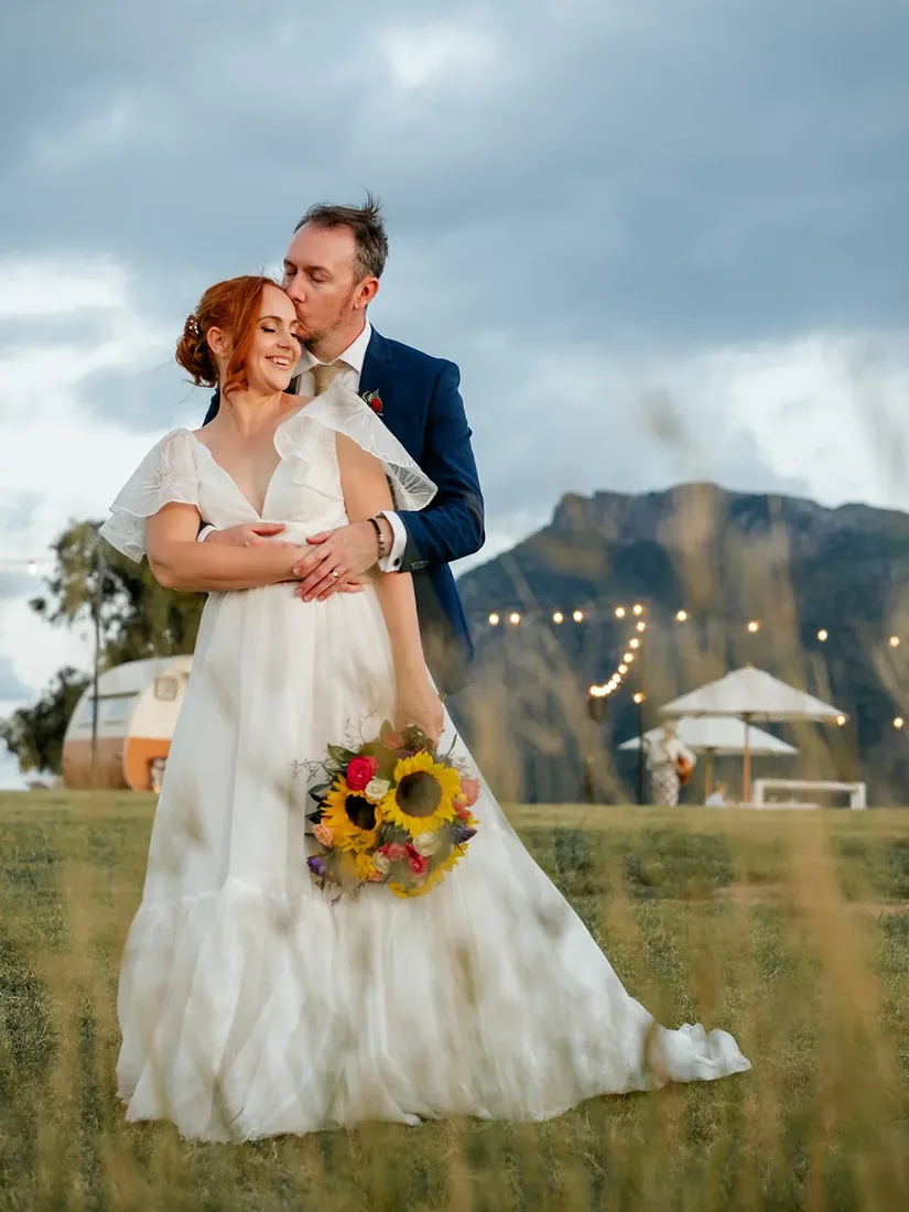 Russell kissing Leisa on the forehead in a golden field at dusk, market umbrellas and string lights behind them