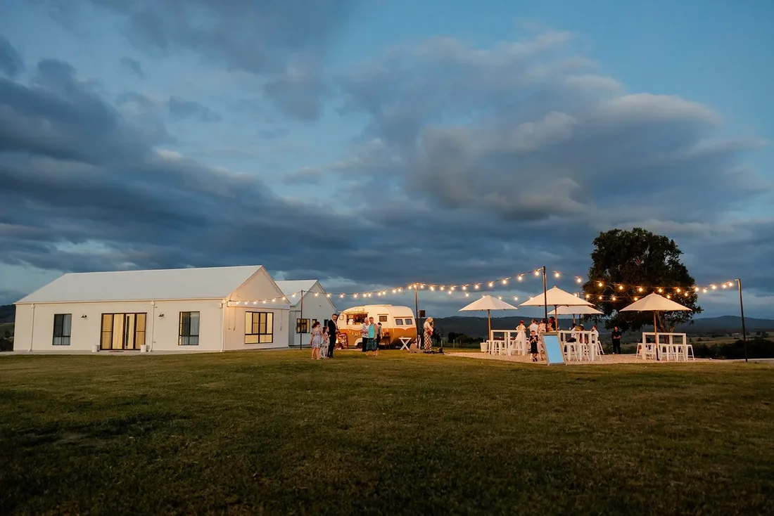 Reception lawn at dusk with festoon lighting, market umbrellas and a vintage caravan bar
