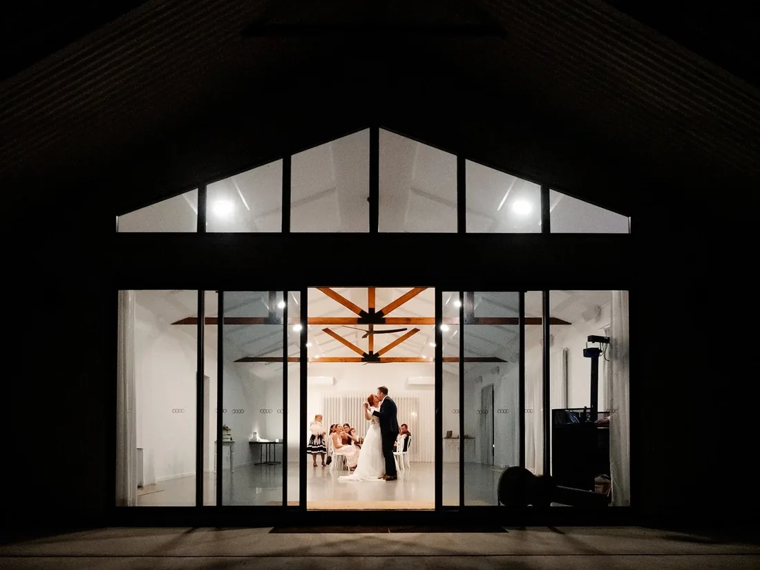 Second view of the reception venue lit up at night, Leisa and Russell dancing inside visible through the glass