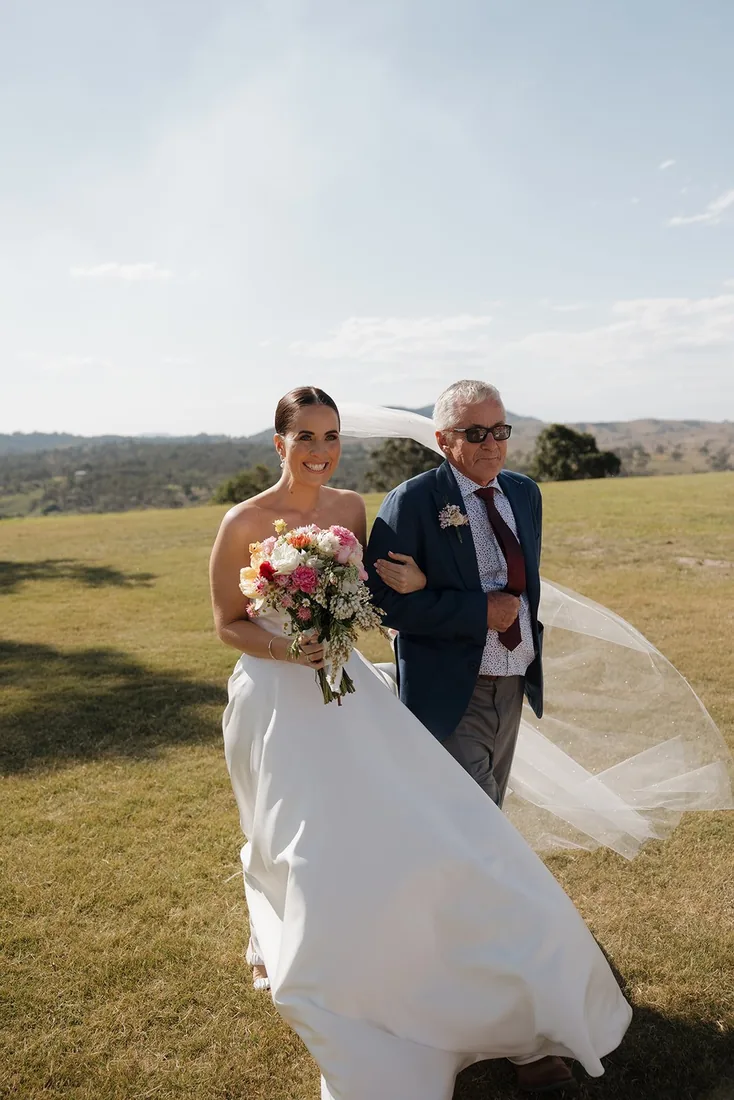Megan walking with her father across the grass, her veil blowing in the wind