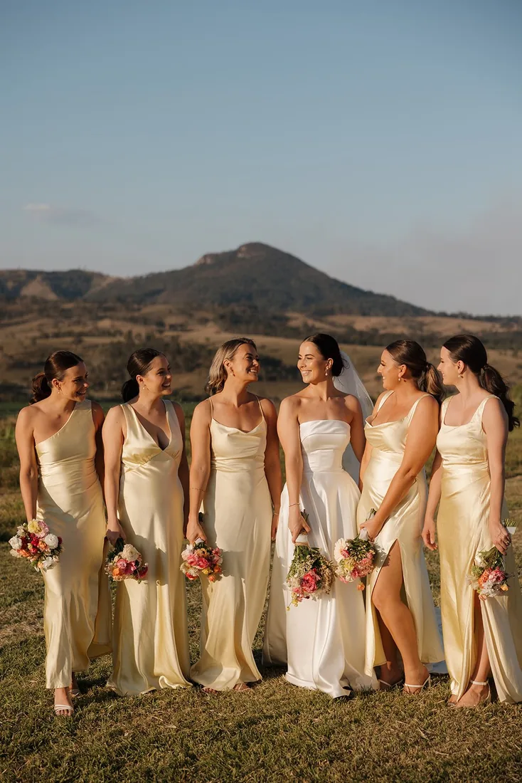 Bridesmaids in matching champagne satin dresses laughing together with Mount Maroon behind them