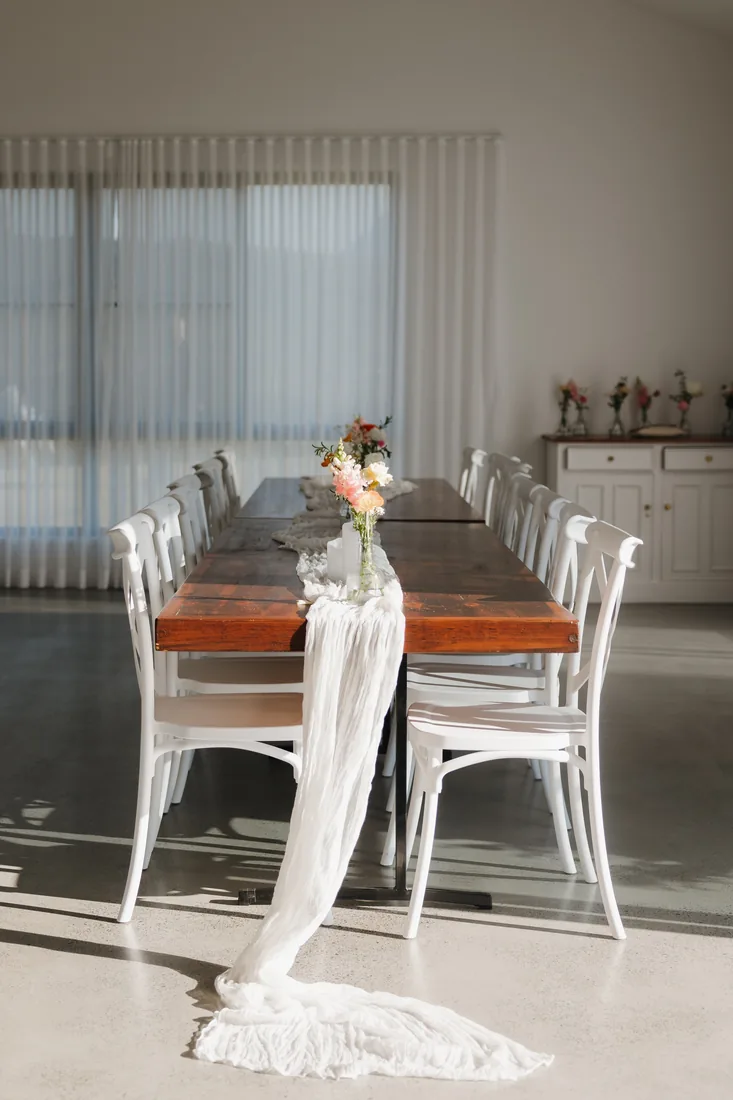 Long dining table set with a timber table, floral runner and white chairs indoors