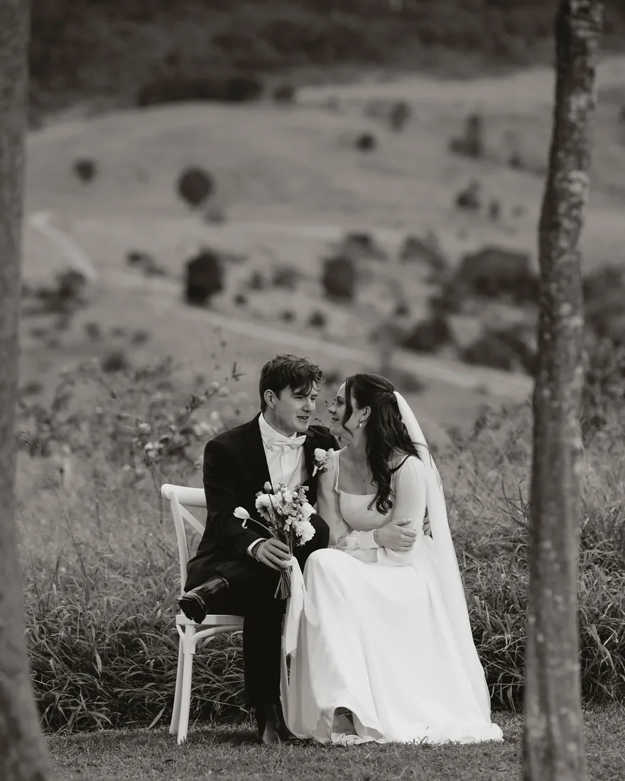 Black and white photo of Melanie and Nathan sitting together, Nathan holding the bridal bouquet