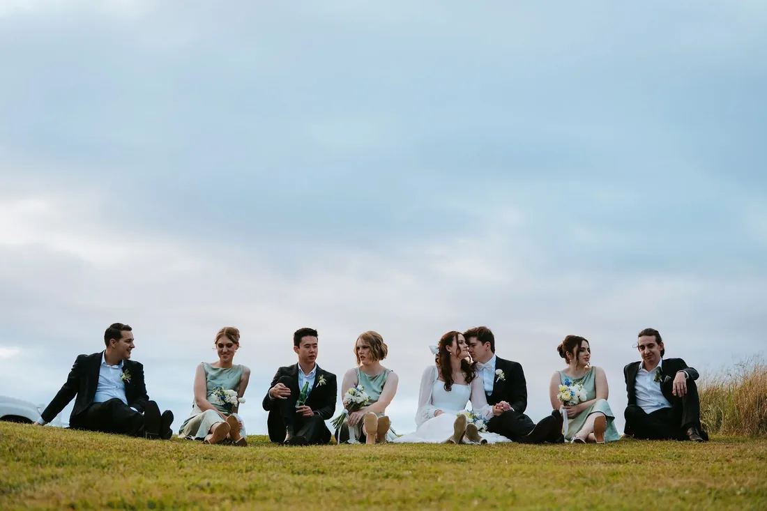 Bridal party sitting together on the grass, laughing, under an overcast sky