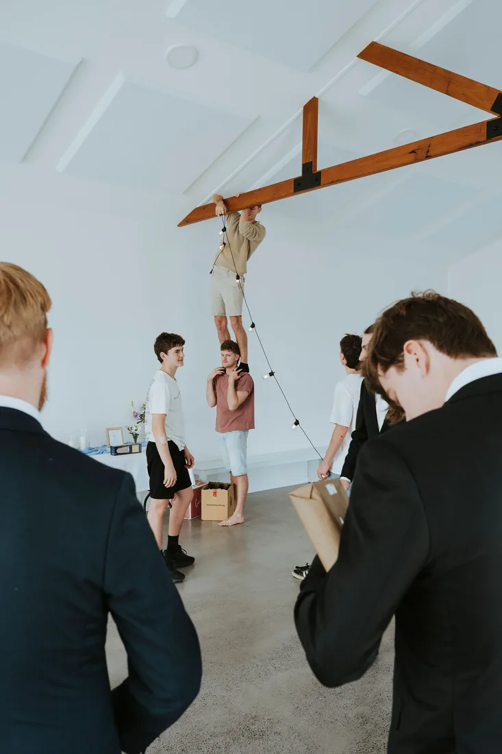 Groomsmen hanging festoon lights from the timber beams inside the reception room
