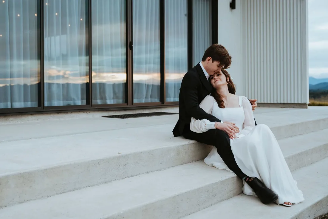 Melanie and Nathan sitting together on the venue steps at dusk, string lights visible in the windows