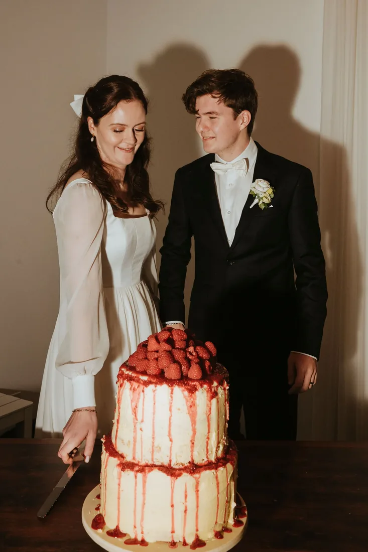 Melanie cutting a two-tiered cake topped with fresh raspberries and a red drip, Nathan beside her