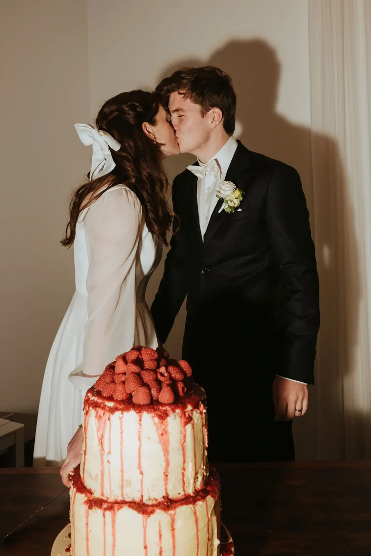 Melanie and Nathan kissing beside their raspberry drip cake at the reception
