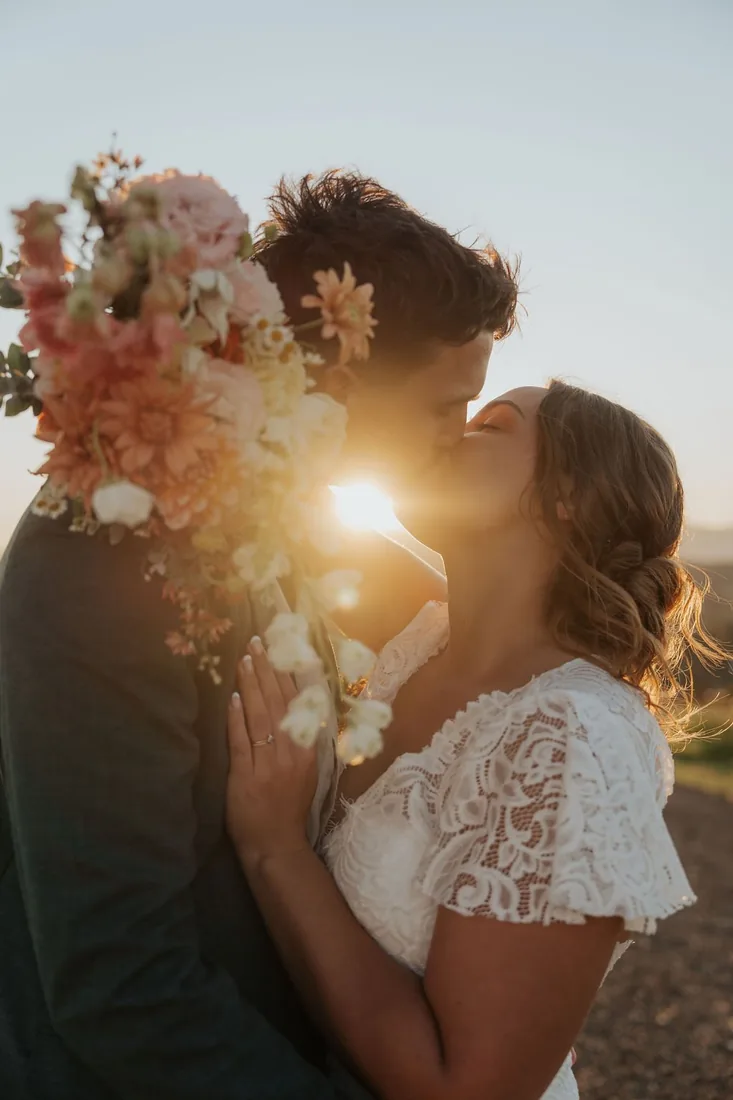 Nicola and Aaron kissing at sunset, golden light behind them and a bridal bouquet between them