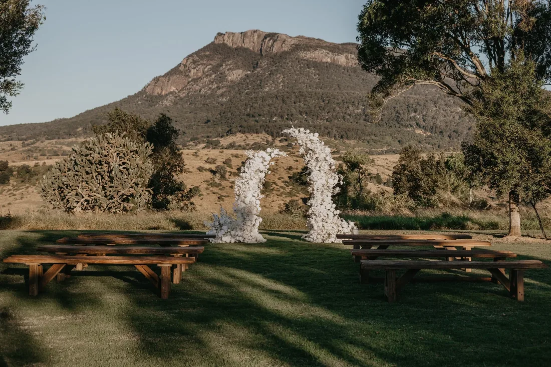 White floral arch ceremony installation framed by rugged Mount Maroon, timber cross-back benches either side