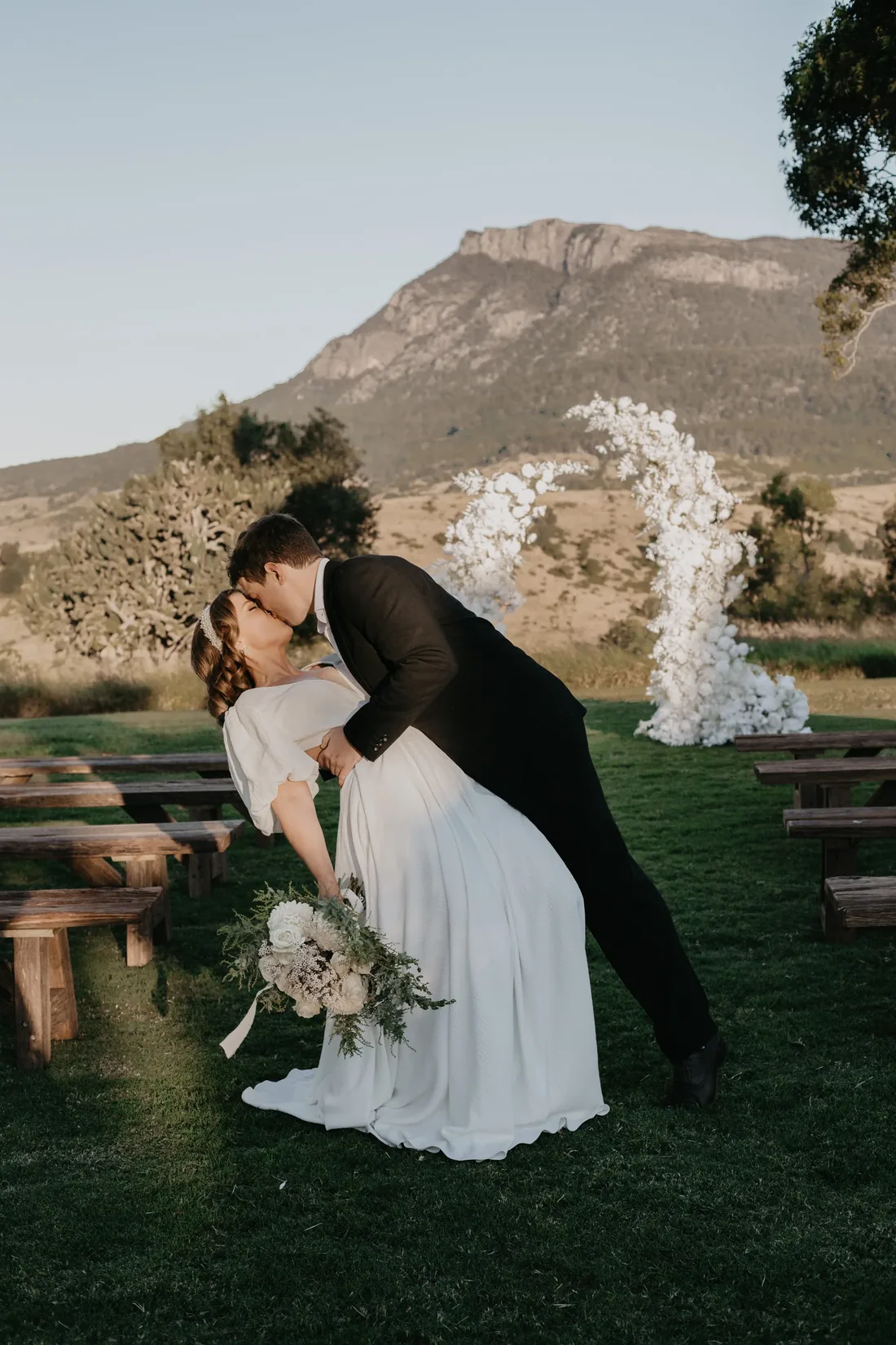 Groom dipping the bride for a kiss beside a white floral arch at Maroon Hill Estate