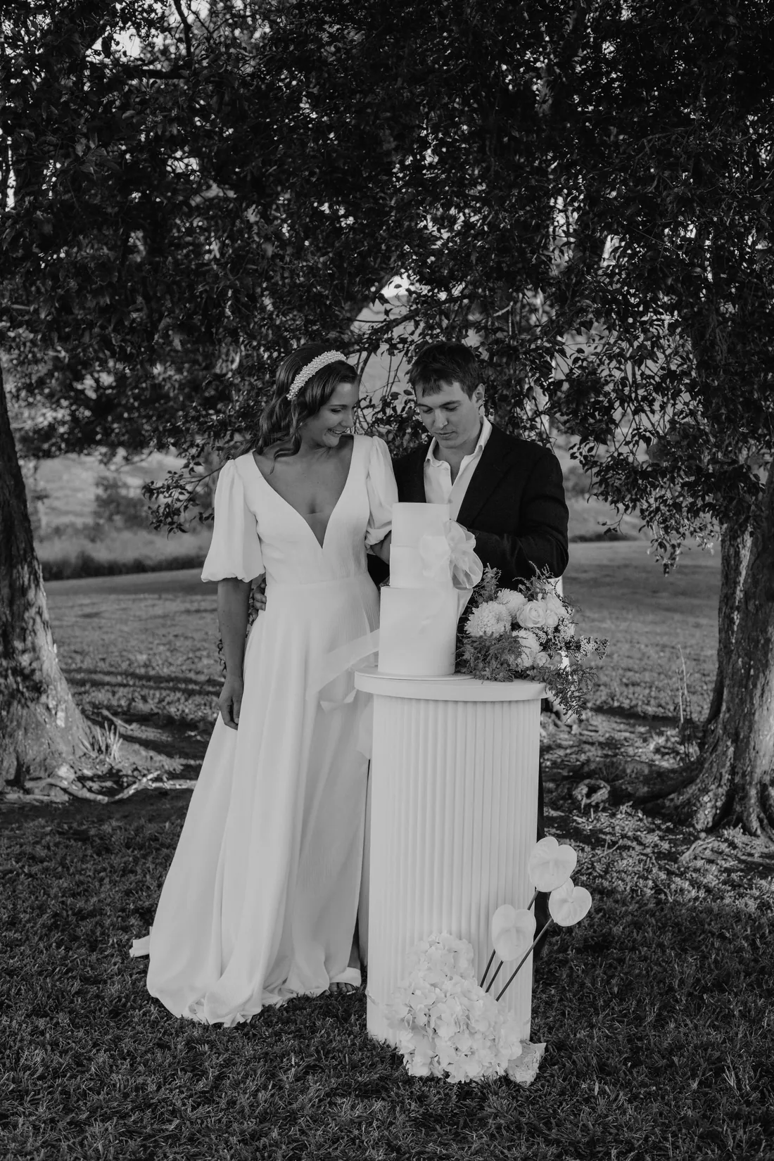 Black and white photo of the couple cutting a two-tiered cake beneath a shade tree