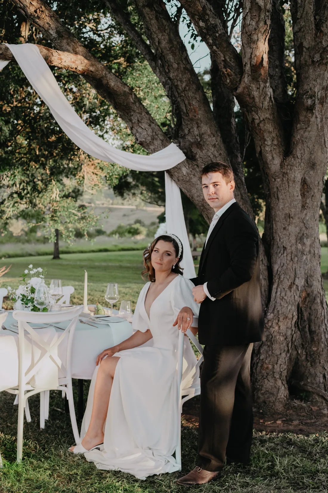 Bride and groom standing beside a styled reception table draped in white beneath a large tree