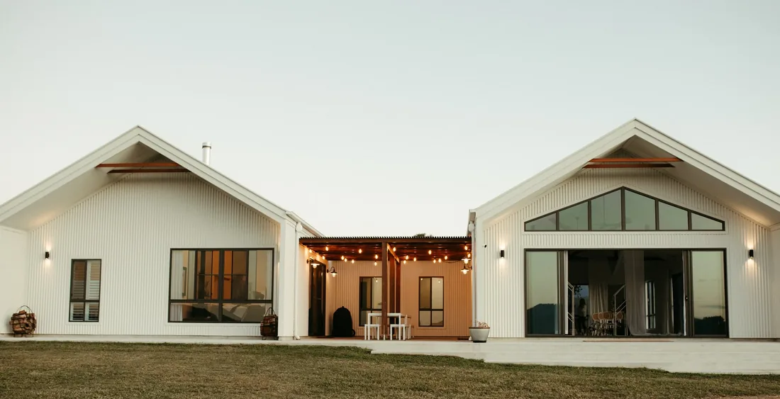 Exterior of the white gable-roofed venue at dusk, string lights glowing on the covered patio between the two barns
