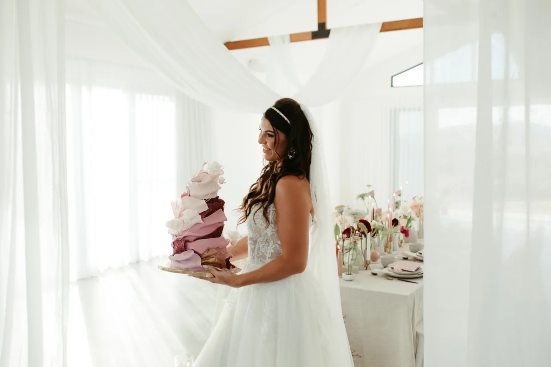 Bride holding a pink and burgundy ombre wedding cake, reception table styled behind her